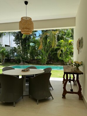 A dining area with a round table and chairs, a hanging light fixture, and a view of a swimming pool and lush greenery.