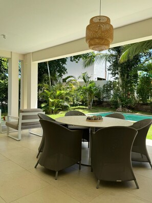 An outdoor dining area with a round table and four chairs, a hanging light fixture, and a view of a swimming pool and greenery. - Villa Robles in Punta Cana (Punta Cana)