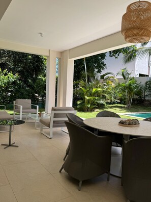 A modern outdoor dining area with a round table and chairs, surrounded by lush greenery and a swimming pool.