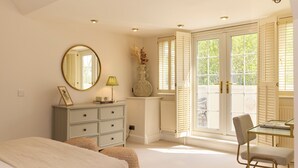 Une chambre à coucher avec un lit, une commode, un miroir, une lampe, un vase et une vue sur l’extérieur, visible à travers une porte.