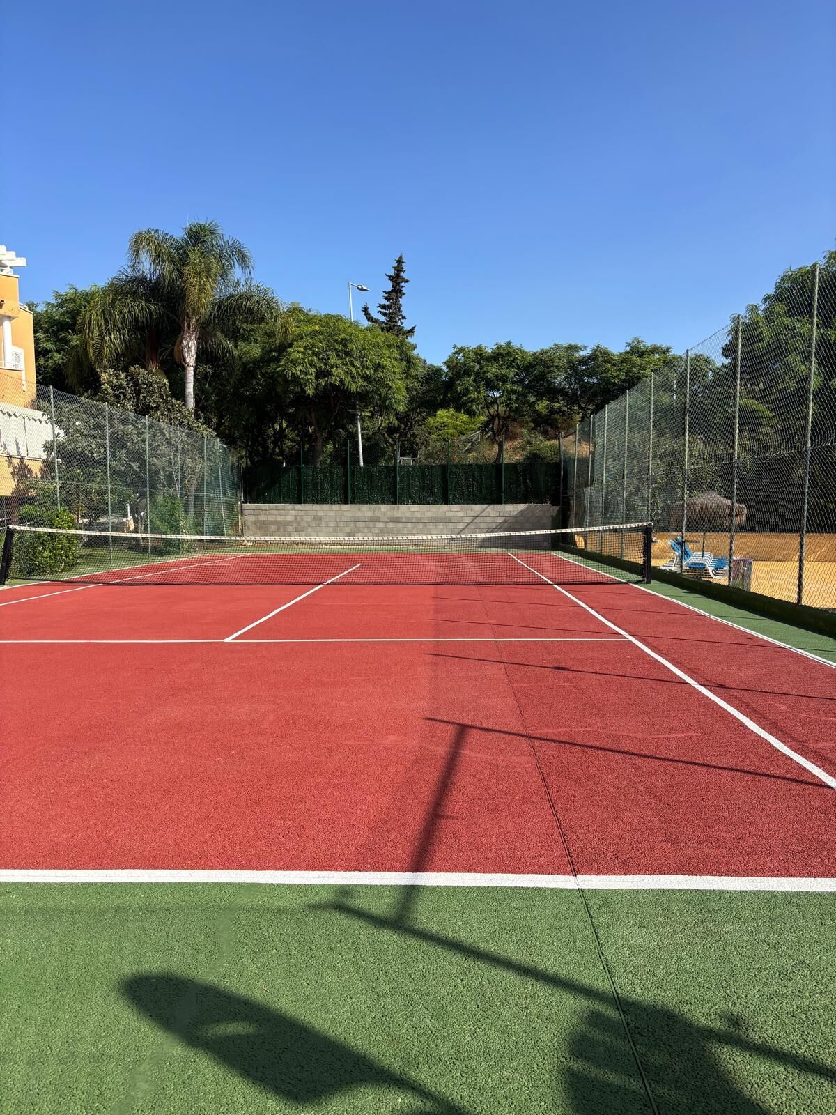 Een tennisbaan met een net, omgeven door een groene hekken en bomen.