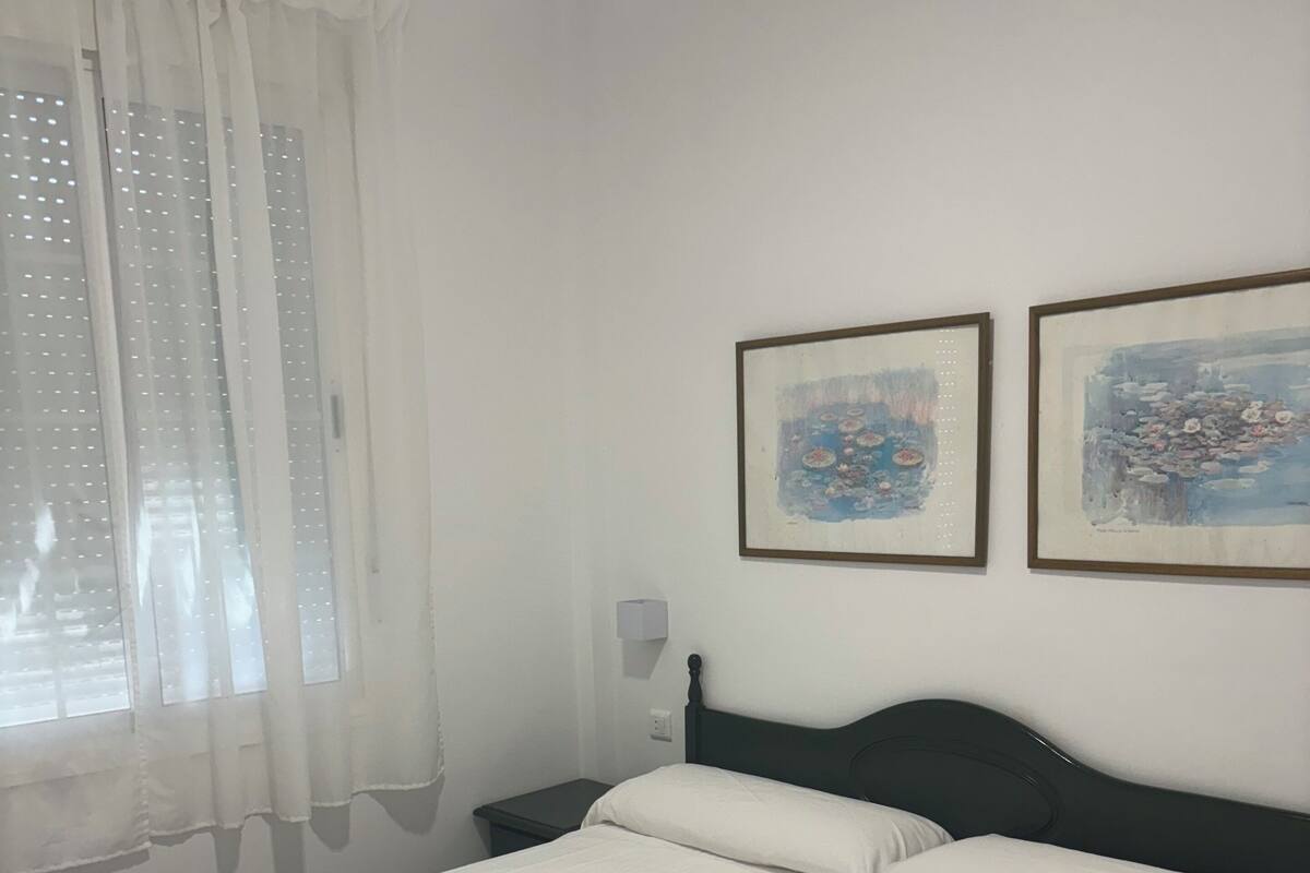 A hotel room with two beds, white bedding, a nightstand, a lamp, and two framed paintings on the wall.