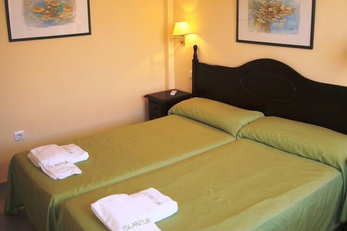 A hotel room with two beds, each with a green bedspread and a white towel labeled "ISLANDUR". There are two framed artworks on the walls and a bedside lamp on the left side.