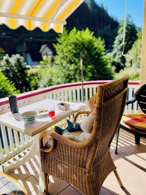 A wicker chair with a table on a balcony, a bottle, and a glass of red liquid. - Ferienhaus Oesterle - Idyllic Black Forest house by a stream with terrace & balcony (Baiersbronn (Schönmünzach))