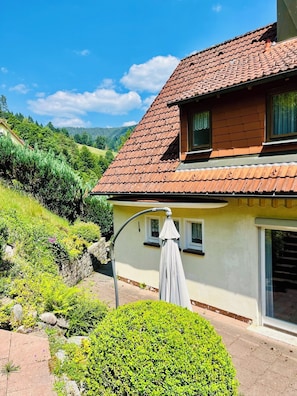 Une maison à deux étages, avec un toit recouvert de tuiles rouges, un petit porche agrémenté d’un parasol blanc et un jardin bien entretenu.