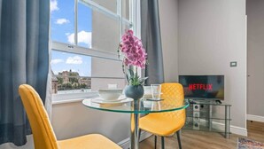 Ein modernes Wohnzimmer mit Glastisch, zwei gelben Sessel, einer Topfpflanzen-Orchidee, einem Netflix-Fernseher und Blick auf Gebäude durch große Fenster.