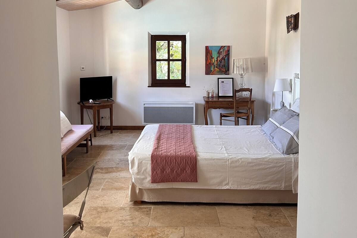 Een slaapkamer met een bed, bureau, stoel, televisie en raam.
