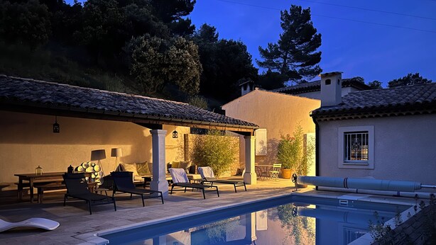 Un área de piscina con sillones de descanso y una pérgola, rodeada por un edificio con tejado de tejas y una ventana pequeña.