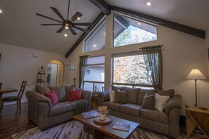 A living room with two sofas, a coffee table, a ceiling fan, and large windows with curtains.