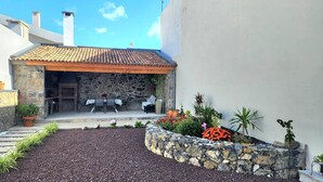 Un edificio de piedra con techo de tejas, un patio con mobiliario exterior y un jardín con diversas plantas y flores.
