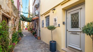 A narrow street with potted plants lining the path, a building with a sign reading "S. PLAZZA RESIDENCE," and a door with a decorative glass panel.
