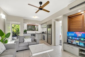 Un salon moderne comprenant un canapé, une table basse, un téléviseur, un ventilateur de plafond, un coin cuisine et un balcon agrémenté de plantes.
