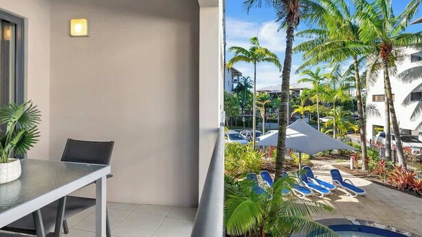Un balcon avec une table et des chaises, un espace piscine avec des chaises longues et des palmiers.