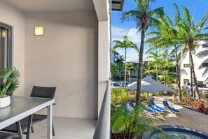 A balcony with a table and chairs, a pool area with lounge chairs, and palm trees. - The Trinity Duo—Resort Comfort near Beach & Dining (Trinity Beach)