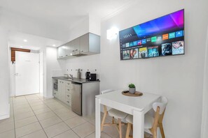Une cuisine moderne équipée d’un téléviseur encastré qui diffuse différentes chaînes, un coin repas avec une table et des chaises blanches, et un couloir menant à une porte.
