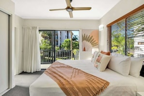 A hotel room with a bed, a ceiling fan, a balcony with a view of buildings and greenery, and a window with blinds.
