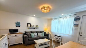 A compact living space with a kitchenette, a grey sofa with decorative pillows, a wicker coffee table, a dining table with a chair, and a ceiling light fixture. - Beach block condo minutes to the boardwalk in Ocean City - Perfect for a getaway (Ocean City)