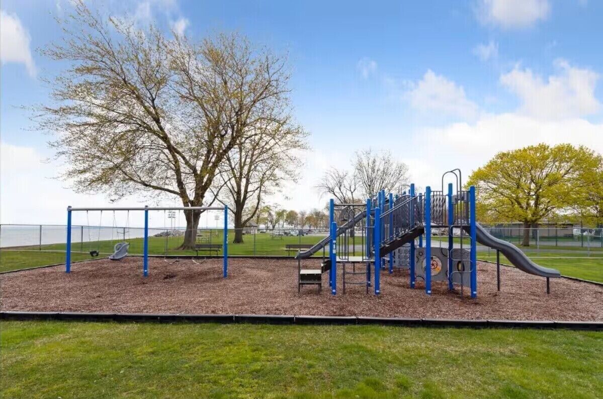 A playground with a slide and climbing structures, surrounded by grass and trees.