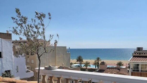 Un balcón con vista al mar, un árbol y edificios al fondo.
