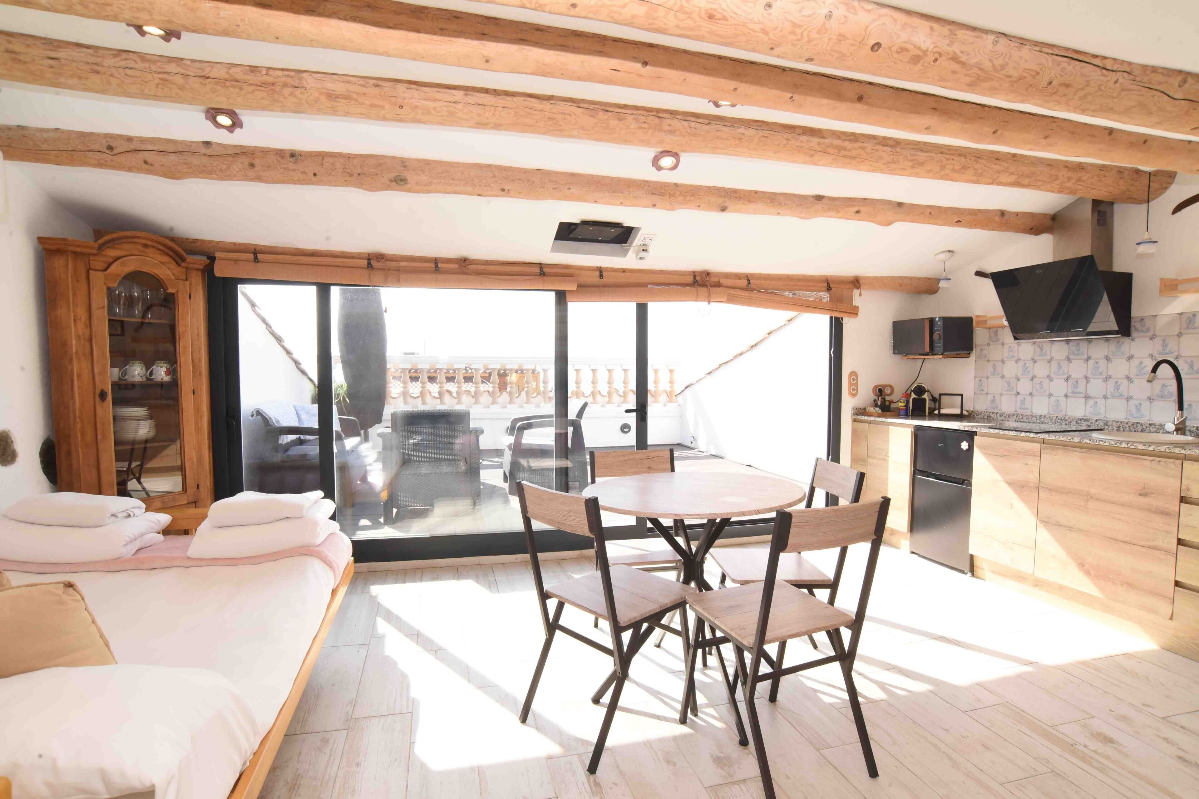 Una cocina moderna con vigas de madera, una zona de comedor con mesa y sillas, horno empotrado y un gran ventanal corredero que da a un patio exterior.