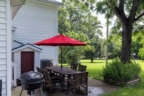 An outdoor patio with a wooden table and chairs, a red umbrella, and a barbecue grill.