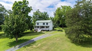A two-story white house with a porch, surrounded by lush green trees and a gravel driveway.