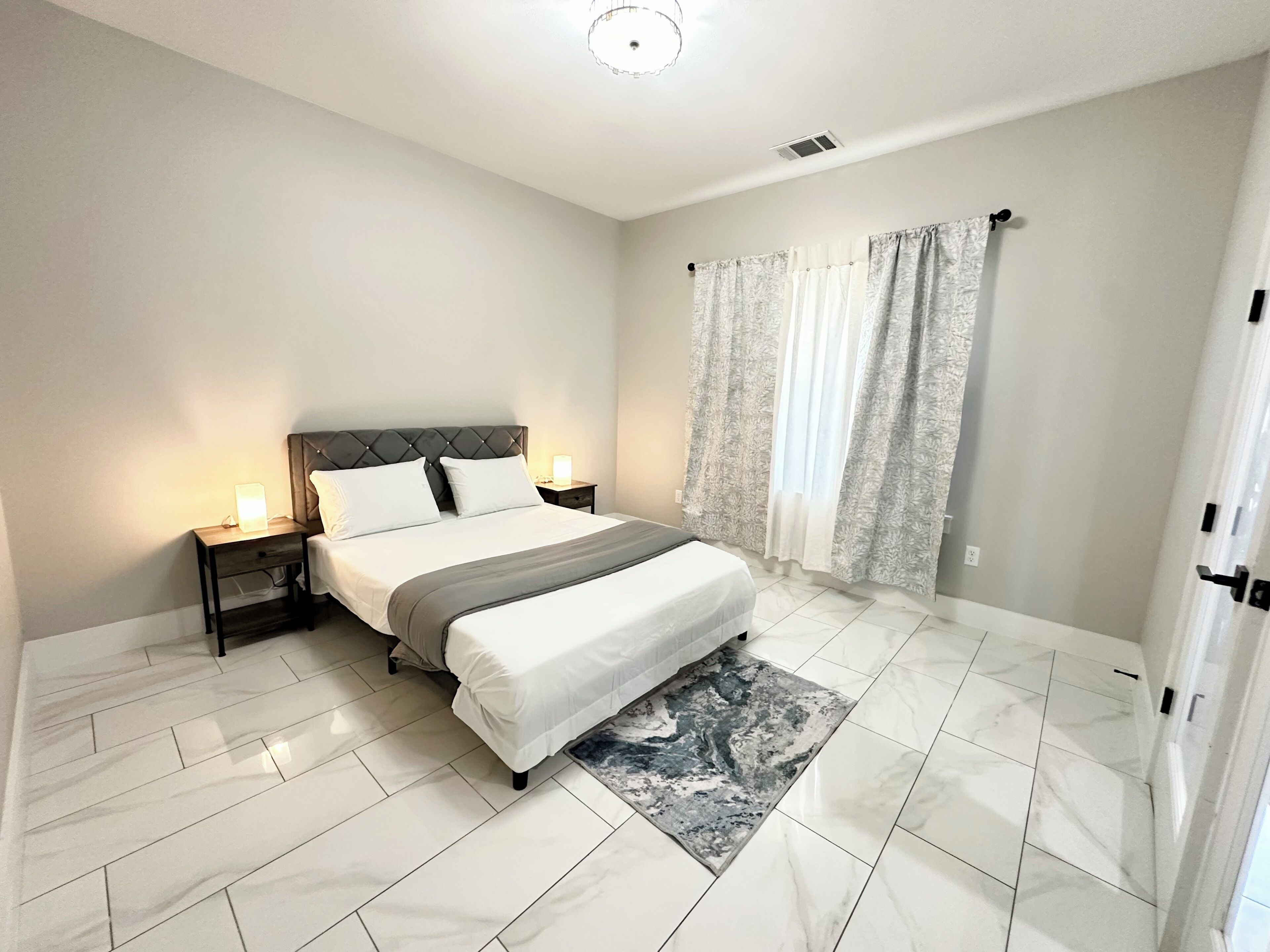 A hotel room with a large bed, two bedside tables with lamps, a window with curtains, and a marble-patterned rug.