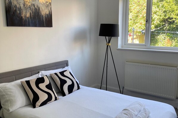 A neatly made bed with white linens and black and white patterned pillows, a floor lamp, and a window with a view of greenery outside.