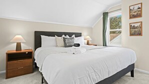 Een net opgemaakt bed met witte lakens, een donker hoofdbord en een kussen met de tekst 'Royal Oak Michigan'. Er zijn twee nachtkastjes met lampen, een houten nachtkastje met lades en een raam met gordijnen.