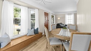 Een eetgedeelte met een ronde houten tafel, witte gordijnen en een bank met kussens.