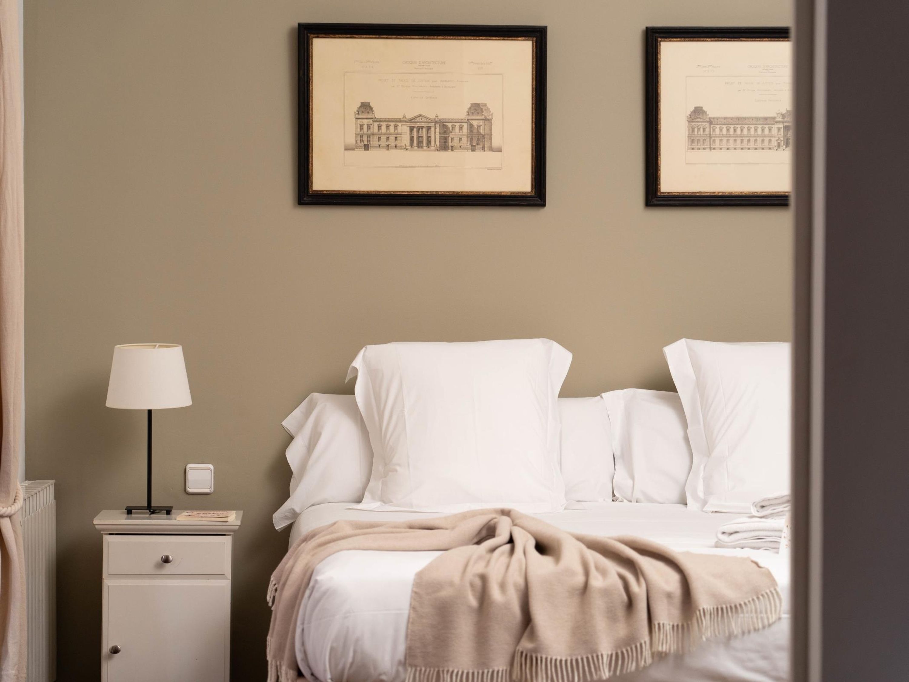 A hotel room with a bed, a nightstand, a lamp, and two framed architectural drawings on the wall.