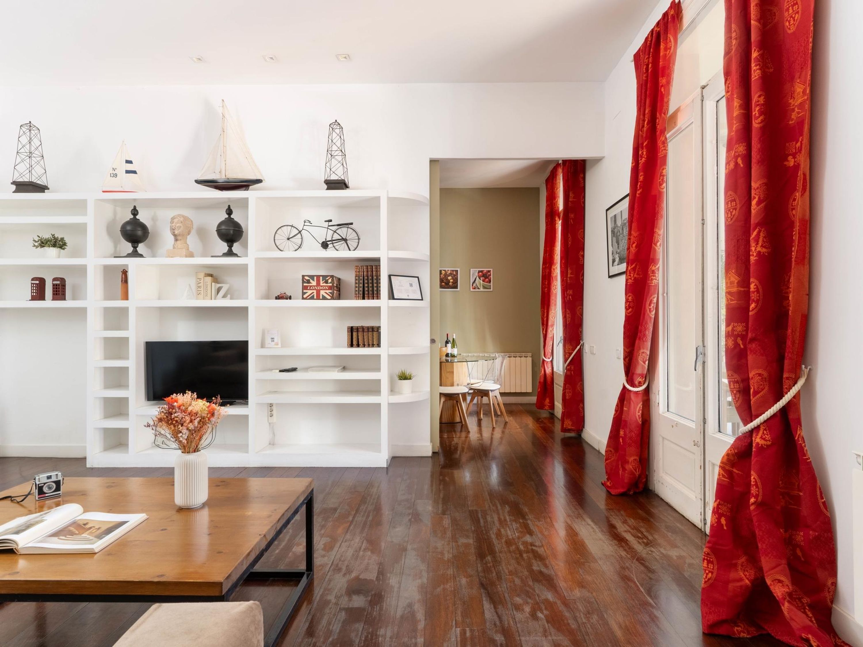 Un salon moderne avec un plancher en bois, des étagères blanches, un coin repas comprenant une table et des chaises, et des rideaux rouges.