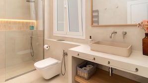 A modern bathroom with a glass shower enclosure, a toilet, a sink with a mirror above, and a small basket under the sink. - Apartamento de Tres Habitaciones con Terraza 11B (Barcelona)