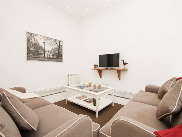 A modern living room with a white sofa set, a coffee table with books and decorative items, a mounted TV, and a framed cityscape picture on the wall. - Apartamento Grande de dos Habitaciones (Barcelona)