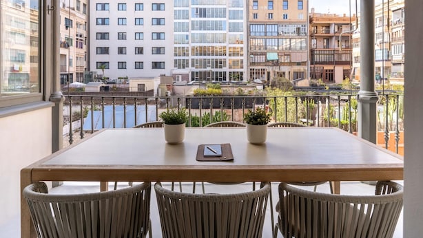 Un balcón con mesa y sillas, con vistas a un paisaje urbano que incluye edificios y una masa de agua.