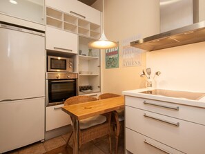 Una cocina compacta con muebles blancos, un microondas, un horno, una mesa de madera, sillas de mimbre y una lámpara colgante.
