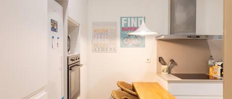 Una cocina compacta con muebles blancos, horno empotrado, extractor de acero inoxidable, una silla de mimbre, una mesa de madera y un aplique de pared.