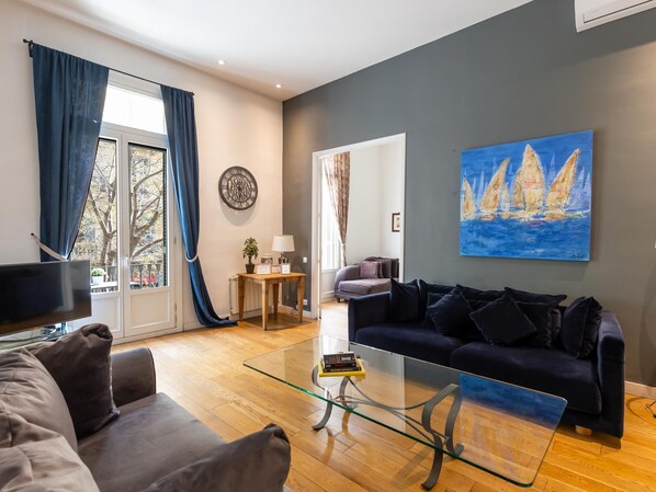 A modern living room with a dark blue sofa, a glass coffee table, and a painting of sailboats. - Apartamento Deluxe de dos Habitaciones (Barcelona)
