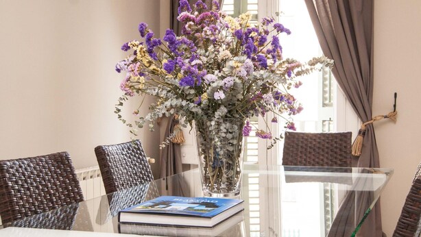A dining area with a glass table, wicker chairs, a vase with flowers, and a book on the table.
