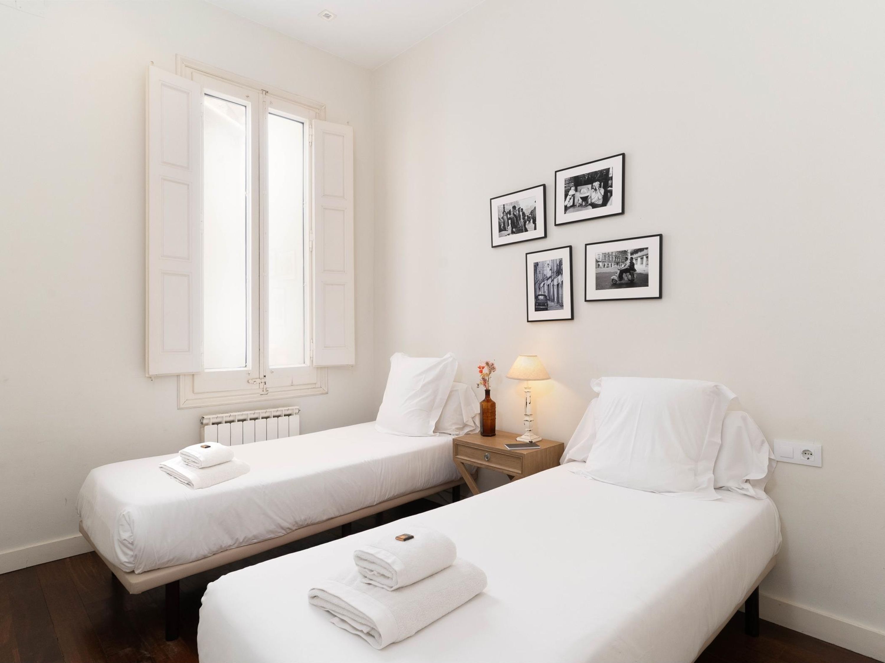 A hotel room with two single beds, a nightstand with a lamp, and a wall with four black-and-white photographs.