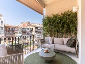A balcony with a sofa, a small table with croissants, and a view of buildings and a pool. - Apto Superior de Tres Habitaciones con Terraza (Barcelona)