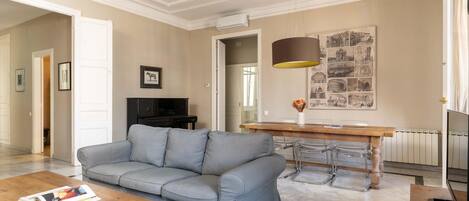 A modern living room with a grey sofa, a wooden coffee table, a dining table with chairs, and a large wall art piece.