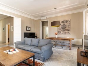 A modern living room with a grey sofa, a wooden coffee table, a dining table with chairs, and a large wall art piece. - Apto Superior de Tres Habitaciones con Terraza (Barcelona)