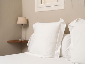 Una cama bien hecha con ropa de cama y almohadas blancas, una mesita de noche con una lámpara y un cuadro en la pared.