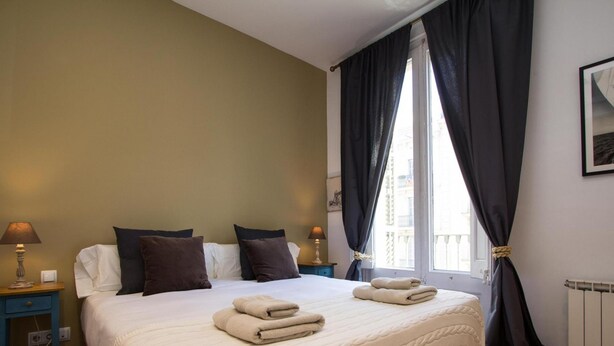 A bedroom with a large bed, two bedside tables with lamps, dark curtains, and a window with a view of buildings. - Apartamento Deluxe de Tres Habitaciones (Barcelona)