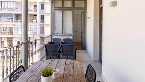 A balcony with a wooden table, two chairs, a potted plant, and a window with blinds. - Apartamento de Tres Habitaciones con Terraza 21B (Barcelona)