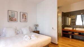 A hotel room with a bed, two framed pictures, a bedside lamp, a wooden bedside table, a bathroom with a large mirror, a sink, and a shower area. - Apartamento de Tres Habitaciones con Terraza 21B (Barcelona)