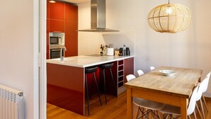 A modern kitchen with a central island, red cabinetry, a stainless steel range hood, and a dining area with a wooden table and white chairs. - Apartamento de Tres Habitaciones con Terraza 21B (Barcelona)