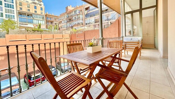A balcony with a wooden table and chairs, a view of buildings, and a car parked below. - Designer s Three Bedroom Apartment With Terrace (Barcelona)