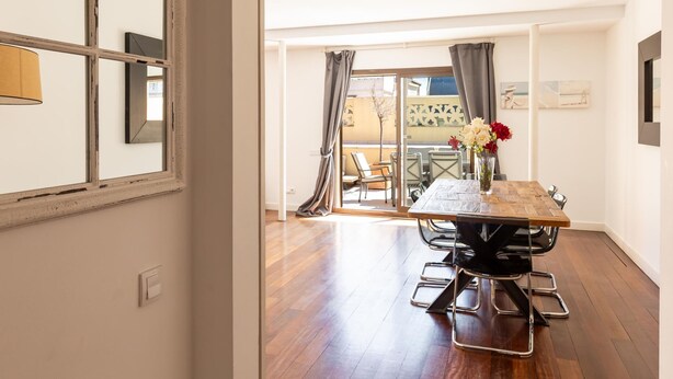 A dining area with a wooden table and chairs, a vase with flowers, and a view of an outdoor balcony with a chair and table. - Apartamento Tico con Terraza (Barcelona)
